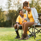 Smiling woman sits in a black Freestyle Rocker as a young soccer player in yellow jersey chats beside her after a game.