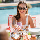 Woman in sunglasses reaches for a snack while seated in a pink floral Freestyle Rocker near a pool.