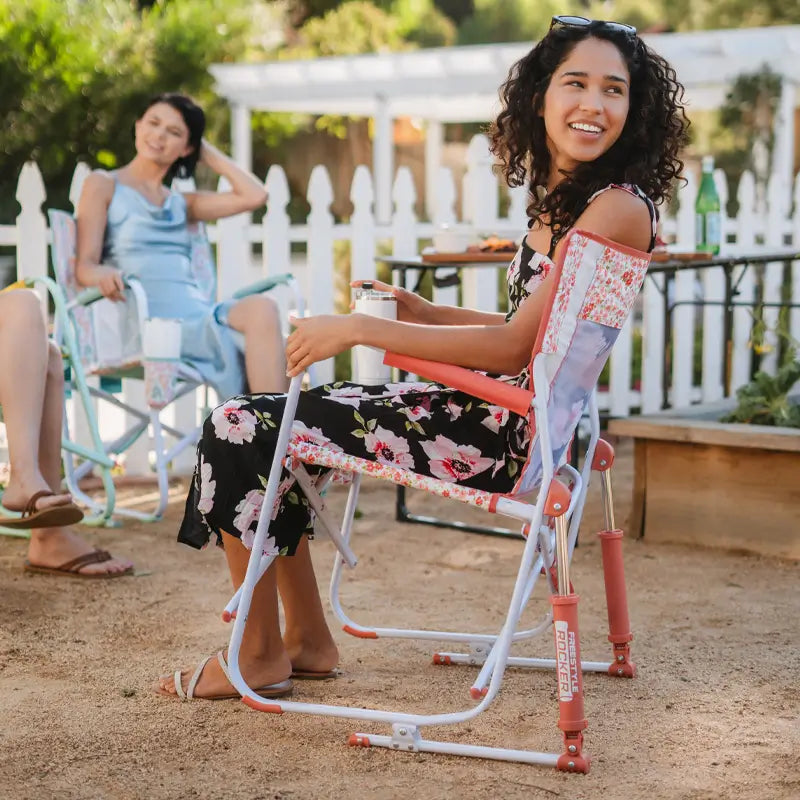 Women enjoy drinks and conversation while sitting in pink floral Freestyle Rockers by a white picket fence.