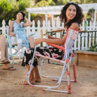 Women enjoy drinks and conversation while sitting in pink floral Freestyle Rockers by a white picket fence.
