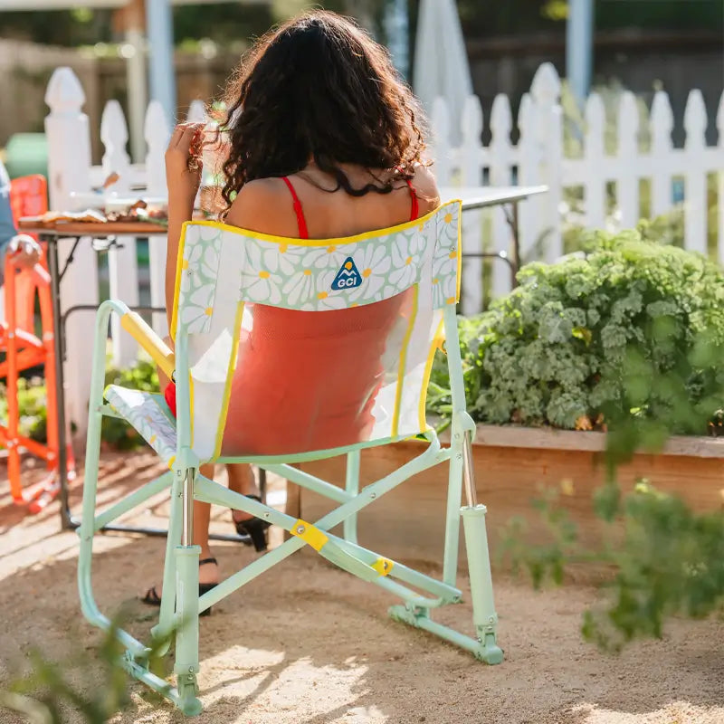 Back view of a woman sitting in the Freestyle Rocker, enjoying a meal at a backyard garden table.