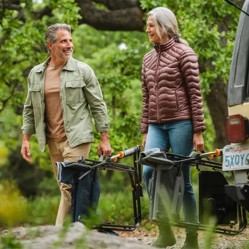 A man and woman smile while carrying folded indigo Freestyle Rocker chairs near a parked vehicle in a forest setting.