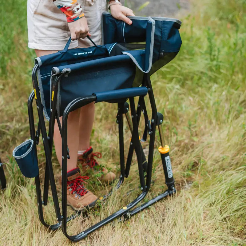 A person in hiking boots stands holding an unfolded navy Freestyle Rocker upright in a grassy field.