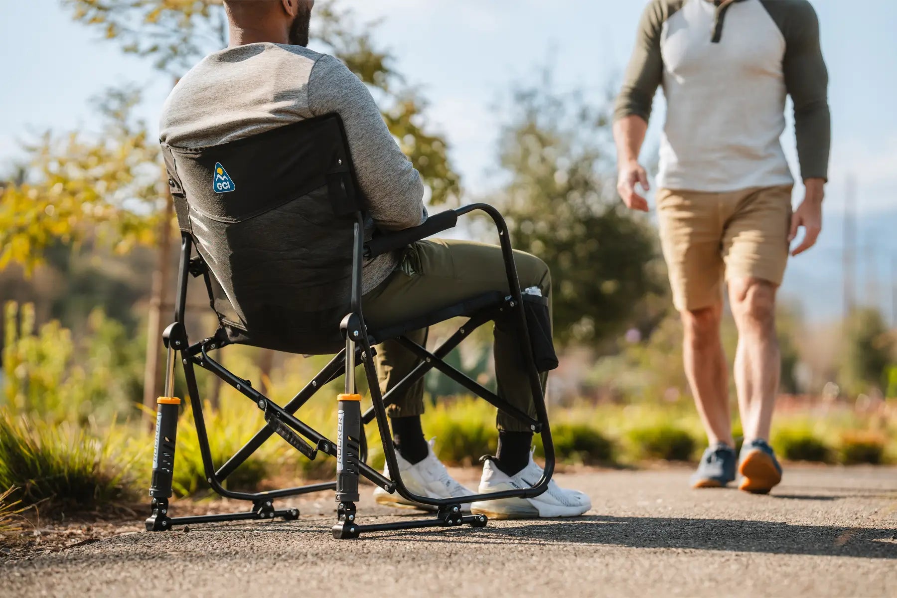 Man relaxes in a Black Freestyle Rocker Elite chair while another person walks by on a sunny outdoor path.