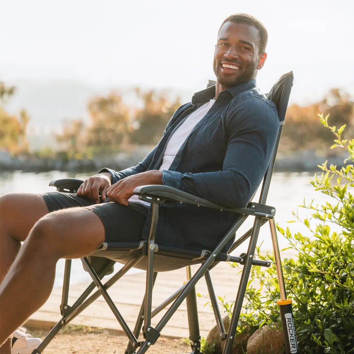 Man smiling and lounging by a lake in a dark-colored GCI Outdoor rocker with Spring-Action Rocking Technology.