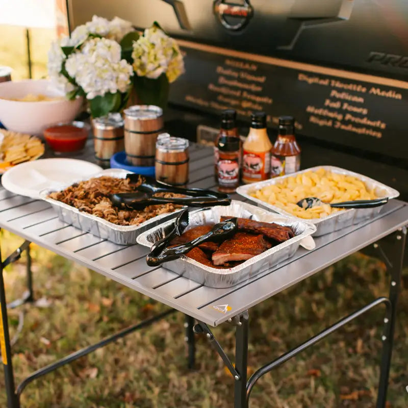 Slim-fold table set with barbecue trays, sauces, and mac and cheese at a tailgate.