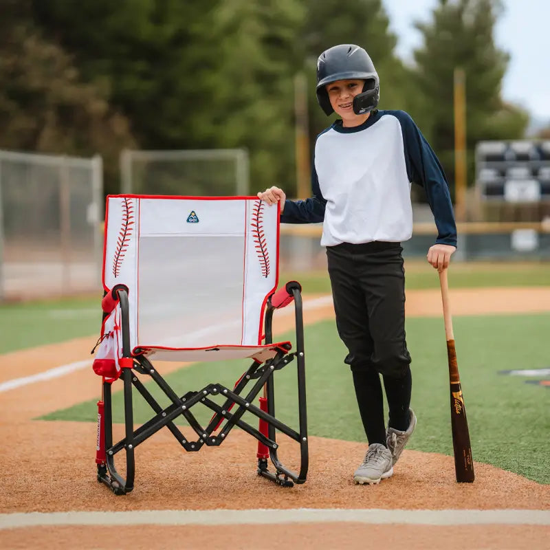A young baseball player in a helmet and uniform stands next to a Stowaway Rocker designed with red baseball stitching, placed near home plate.