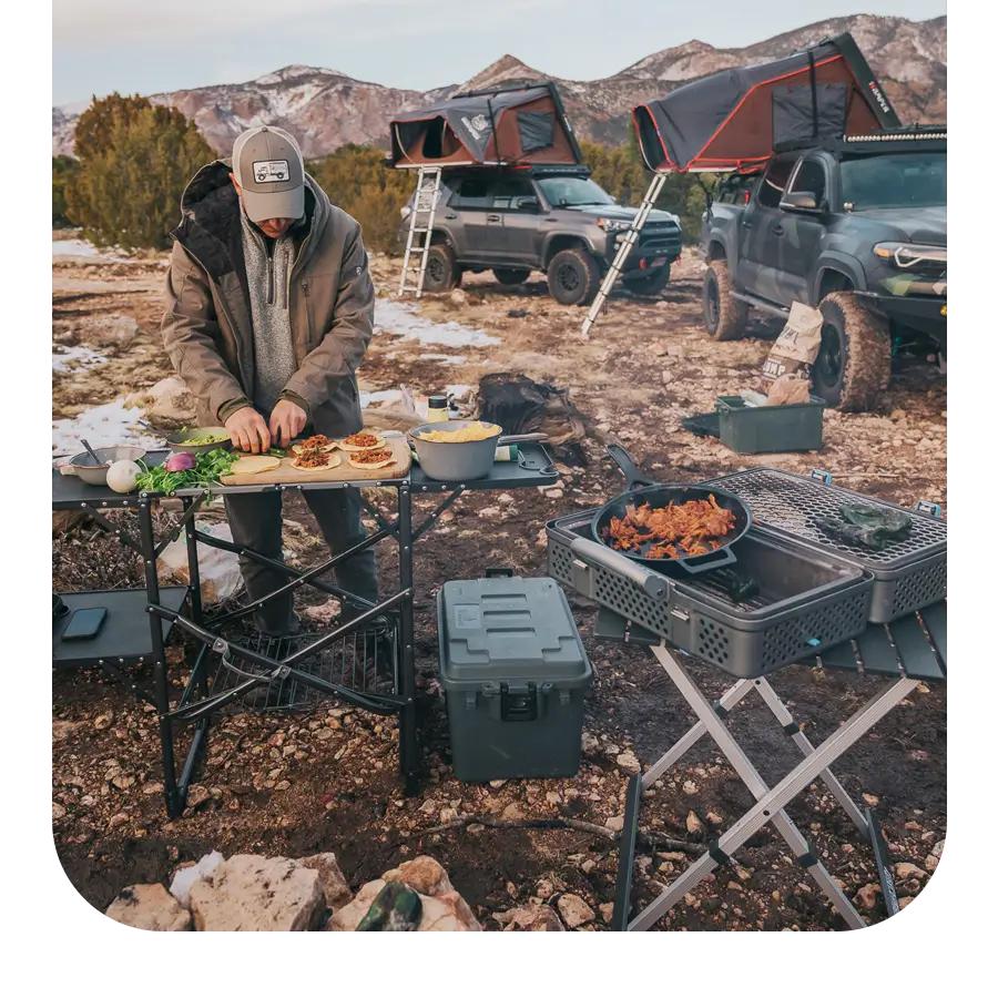 Man assembling tacos on a slim fold cook station near two vehicles with rooftop tents.