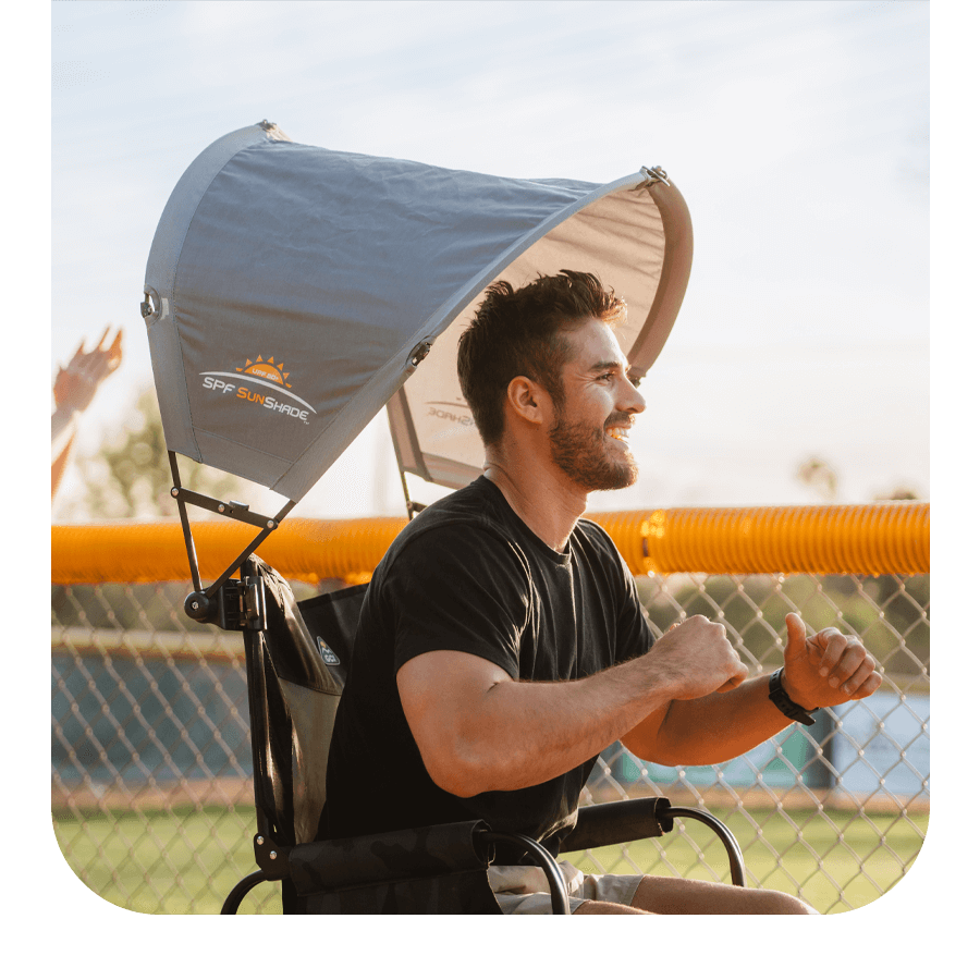 Man smiling while seated in a 168飞艇全国统一开奖结果查询官网 GCI Outdoor camp chair with an overhead SPF SunShade canopy, enjoying a sunny day near a sports field.